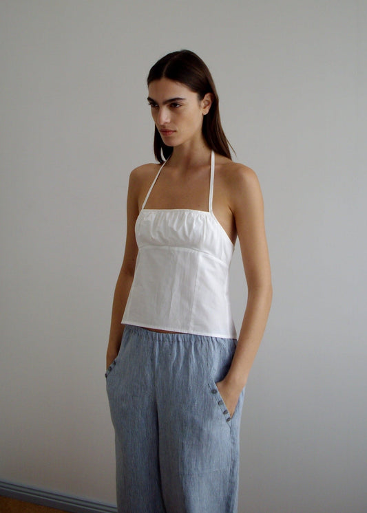 A woman with long brown hair wears the Strap Top - White by Deiji Studios, featuring a gathered bust panel, paired with loose light blue pants. She stands against a plain light-colored wall with her hands in her pockets.