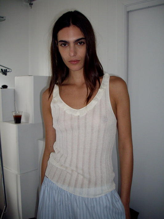 A woman with long dark hair wears the Neck Ruffle Knit in Off White by Deiji Studios and loose, light blue striped pants while standing indoors near a white wall and door. A drink with a straw sits on a ledge behind her.