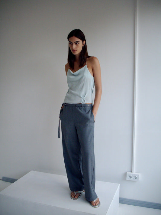 A woman with straight dark hair stands on a white platform against a plain wall, wearing a Deiji Studios light blue sleeveless top, Wool Belt Pant - Grey Blue Stripe, and sandals, with her hands in her pockets.