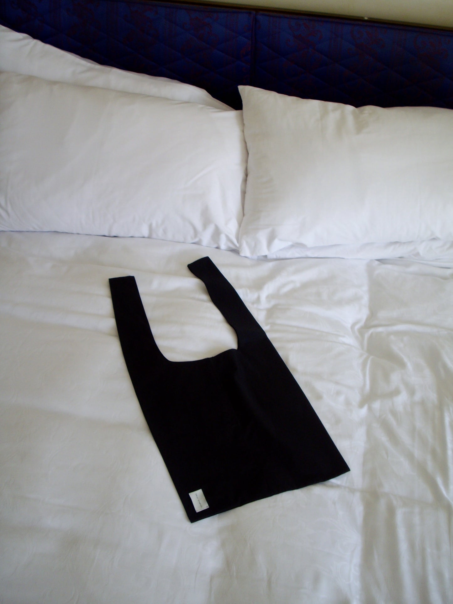 The Seam Tote - Black by Deiji Studios, made from organic cotton, is displayed on a neatly made bed with white sheets and pillows.