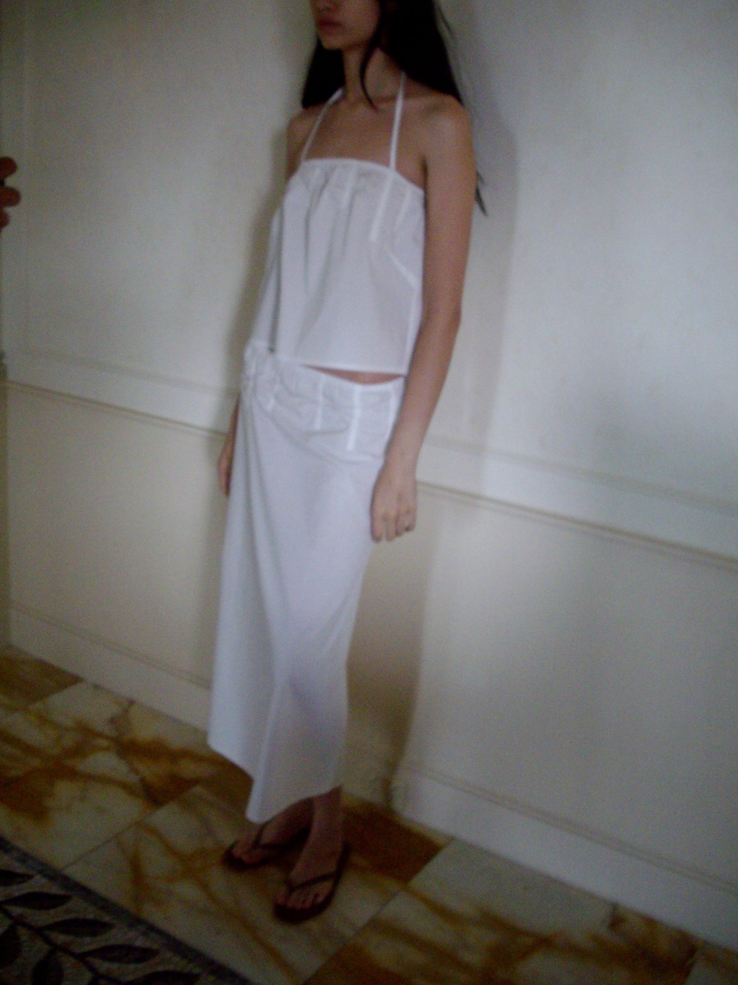 A woman stands indoors against a light wall and marble-patterned floor, wearing the Tube Top - White paired with a matching long skirt and flip-flops. Her hair is loose and her face is partially out of frame.
