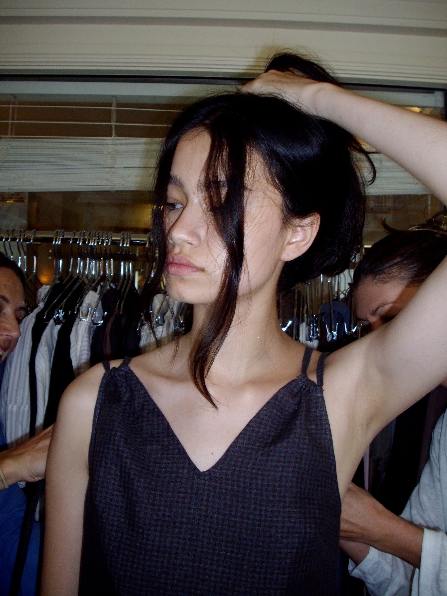 A young woman with long dark hair stands in front of a clothing rack, looking thoughtful as she rests her hand on her head. She wears the Looped Dress - Slate Check, featuring a dark sleeveless design and v neckline.