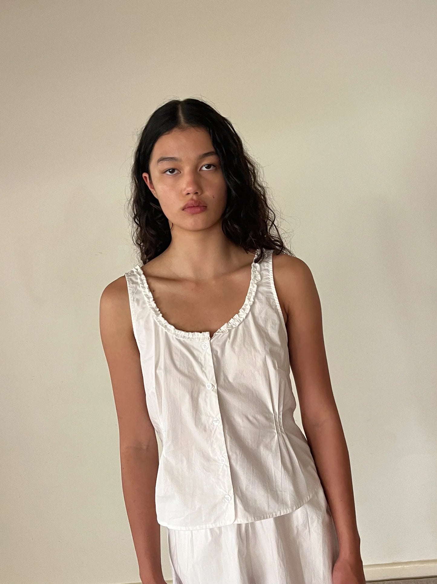 A woman with long wavy dark hair stands against a plain beige wall, wearing the PJ Short Set - White from Deiji Studios. She looks directly at the camera with a neutral expression.