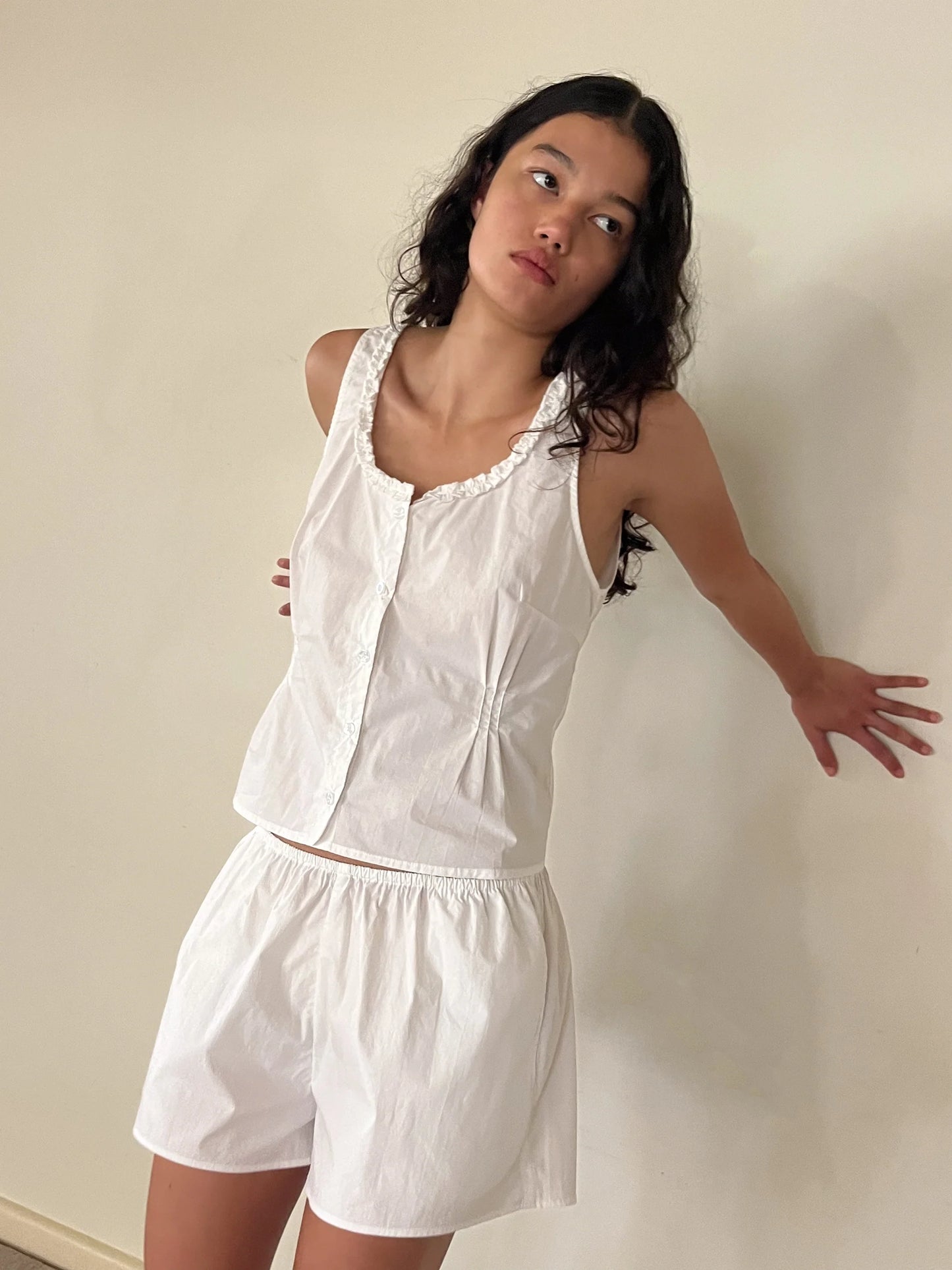 A woman with long dark hair stands against a plain wall, wearing the PJ Short Set - White—sleeveless and loose—looking slightly to the side with a thoughtful expression.