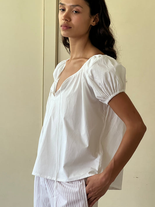 A woman with long dark hair stands sideways against a plain beige background, wearing the One Panel Top—a loose white short-sleeve blouse with a gathered neckline—paired with light striped pants and one hand in her pocket.