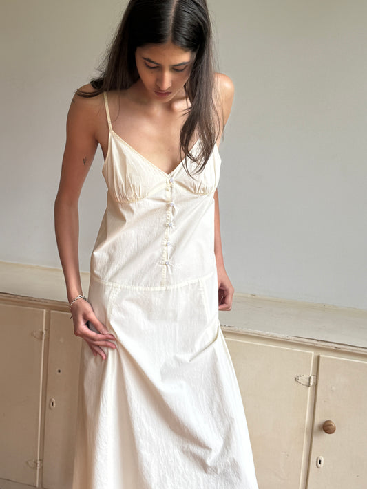 A woman with long dark hair looks down, wearing the Double Seam Dress—a cream-colored, sleeveless design with thin straps and button details—while standing indoors by a light cabinet.