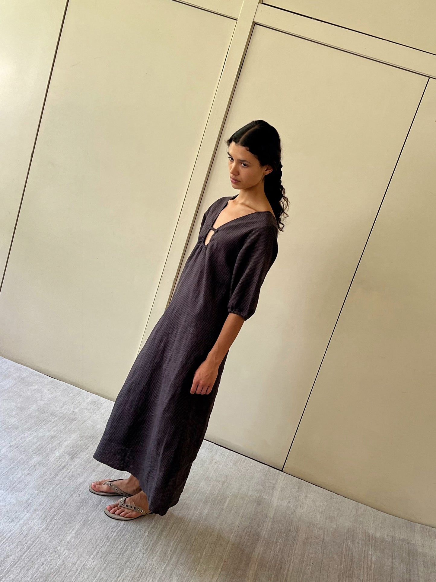 A woman with dark hair, wearing Deiji Studios Squared Dress - Slate Check and sandals, stands indoors against a beige paneled wall, looking slightly to the side.