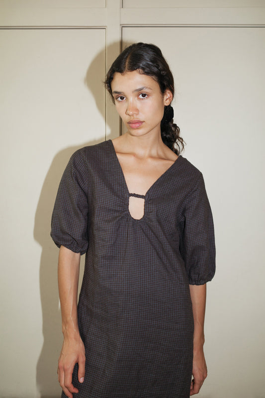 A young woman with long, dark braided hair wears the Squared Dress—dark, loose-fitting with puffed sleeves and a keyhole neckline—standing against a plain cream background.