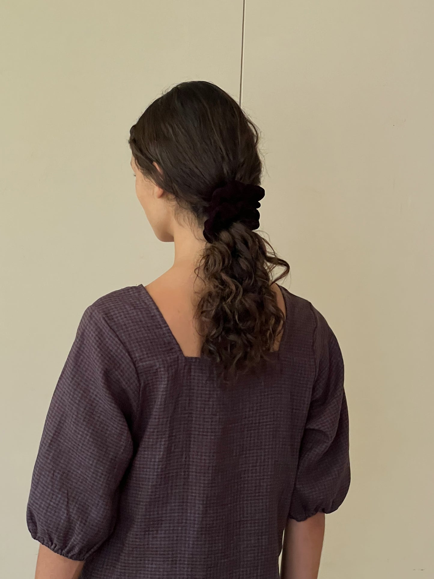 A person with wavy brown hair tied in a ponytail with a black scrunchie stands facing away, wearing the Squared Dress - Slate Check from Deiji Studios. The plain background is beige.