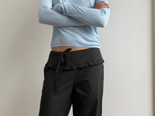 A person stands against a plain light-colored wall, arms crossed, wearing a light blue ribbed long-sleeve top and the Flounce Belt Pant - Black by Deiji Studios, featuring a ruffled waistband and tied belt.