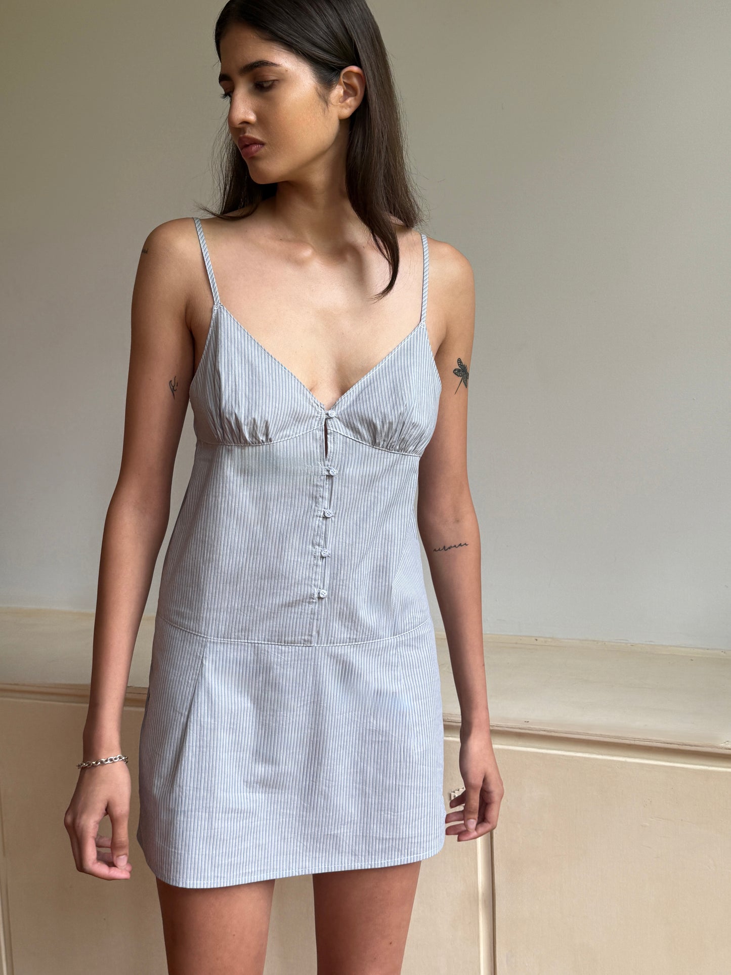 A woman with long dark hair models the Seam Front Dress - Sky Stripe by 0, a mini-length light gray striped organic cotton dress with thin straps, as she stands indoors against a plain background looking to the side.
