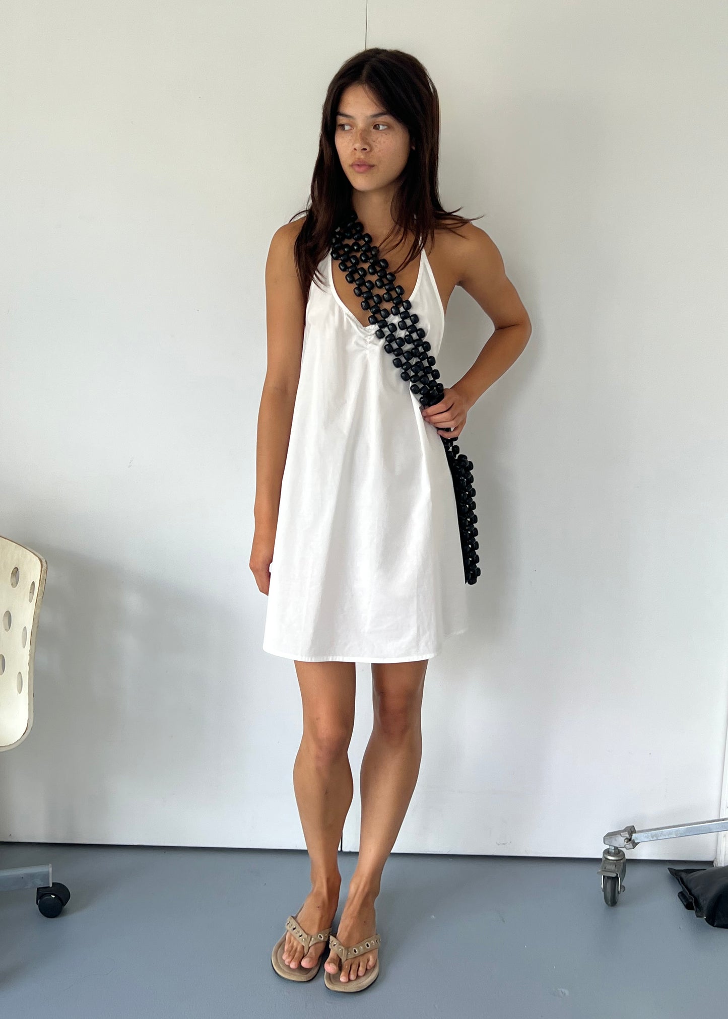 A woman stands indoors against a white wall, wearing the Round Halter Dress - White by Deiji Studios and tan sandals. She has straight dark hair and a chunky black chain strap slung diagonally across her body.