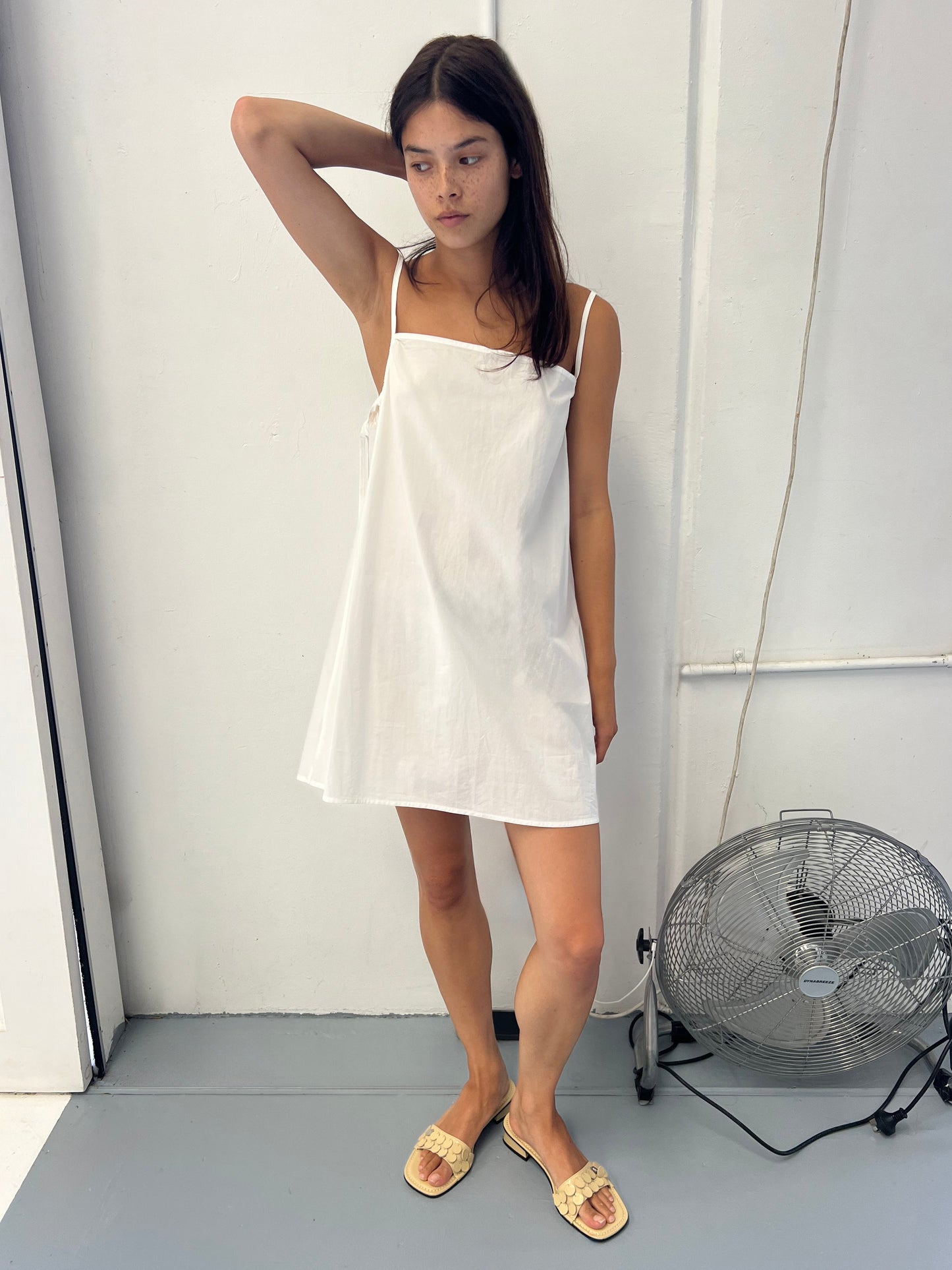 A young woman stands indoors near a wall and floor fan, wearing the Drawcurve Dress—a loose, sleeveless white dress—and woven sandals, with one hand resting on her head and a neutral expression.