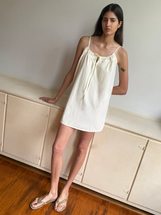 A woman with long dark hair, standing indoors by a light cabinet, wears the sleeveless Light Dress in off-white with clear sandals. A small bird tattoo is visible on her upper left arm.