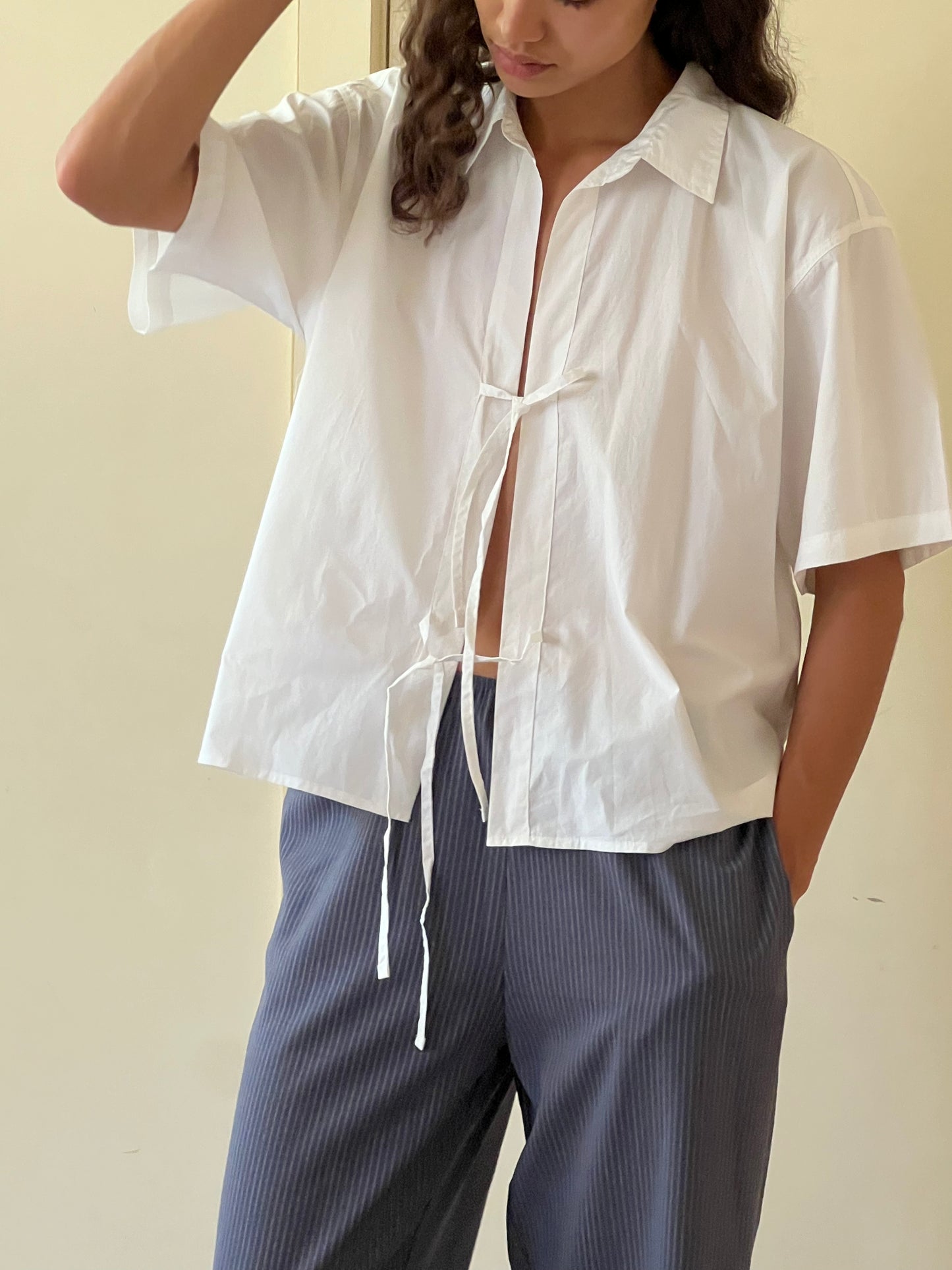 A person wears The Tie Up Shirt, a loose white short-sleeve top tied at the chest, styled with blue pinstripe pants and standing with one hand in their pocket against a plain background.