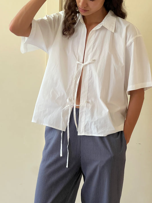 A person wearing The Tie Up Shirt—a loose white short-sleeve shirt with front ties—paired with blue pinstripe pants, stands with one hand in the pocket and the other touching their head.