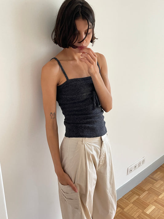 A person with short dark hair wears a dark sleeveless top and the Gathered Pocket Pant in light beige, standing indoors against a white wall with one hand in their pocket and the other near their face.