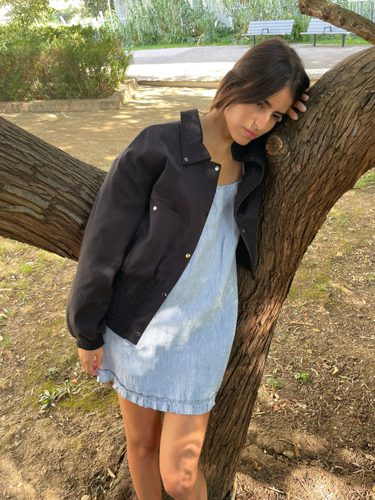 A woman in a light blue dress and a Shirring Jacket - Slate leans against a tree branch in a park, looking down thoughtfully as sunlight filters through the trees onto the ground.
