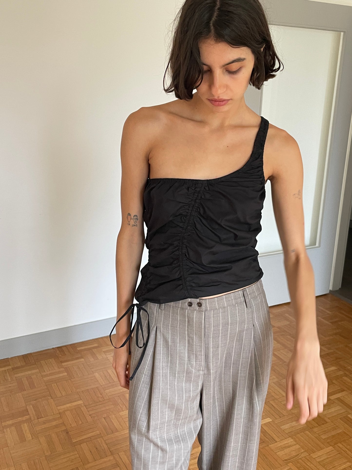 A woman with shoulder-length dark hair wears the Scrunchie Top - Black from Deiji Studios and light gray pinstriped trousers, standing indoors on a wooden floor, looking down with one arm relaxed by her side.