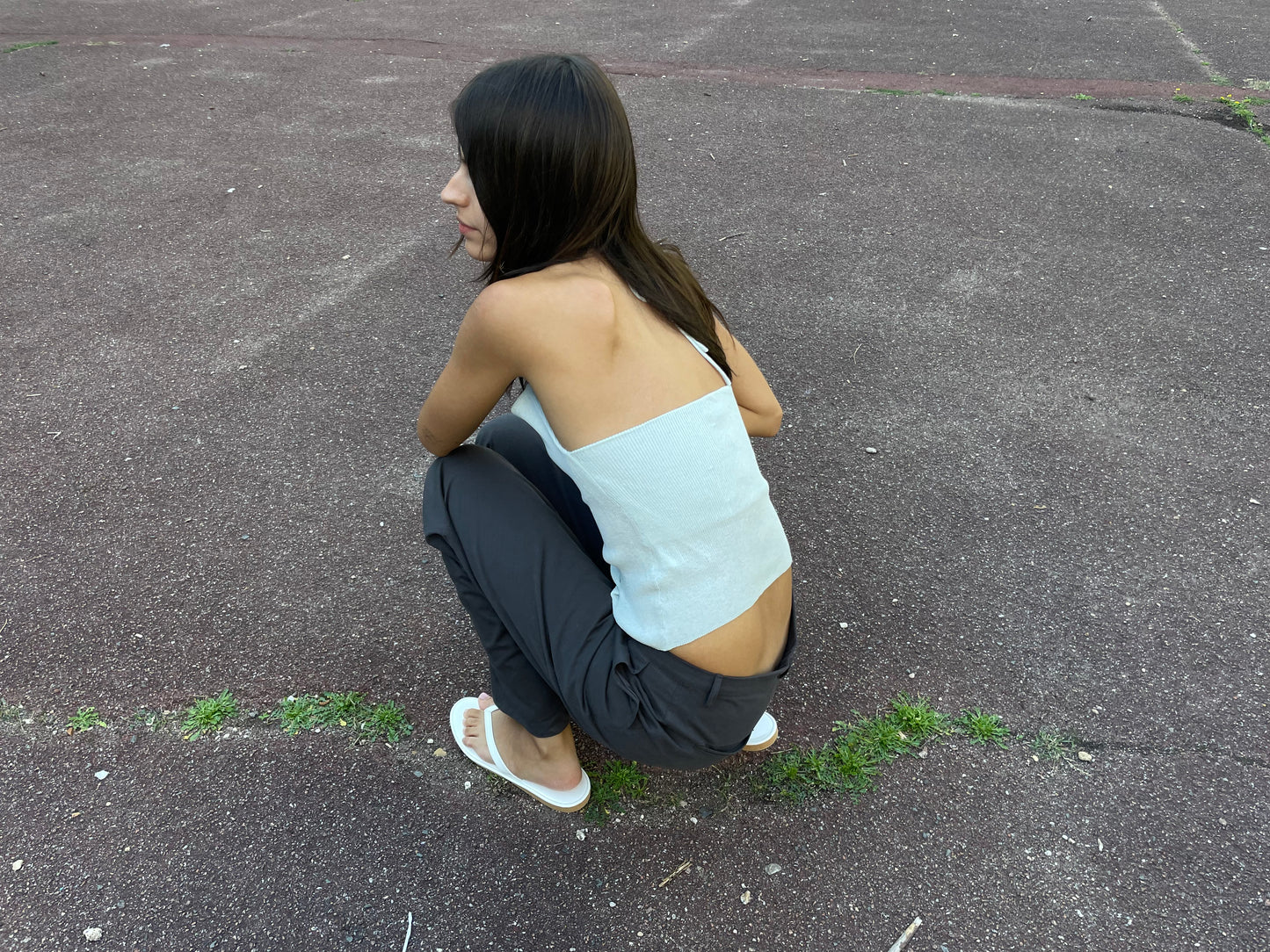 A person with long dark hair squats outdoors on asphalt, wearing a light blue one-shoulder top and the Deiji Studios Zip Pant - Charcoal. White sandals complete the look as grass grows through cracks in the pavement.