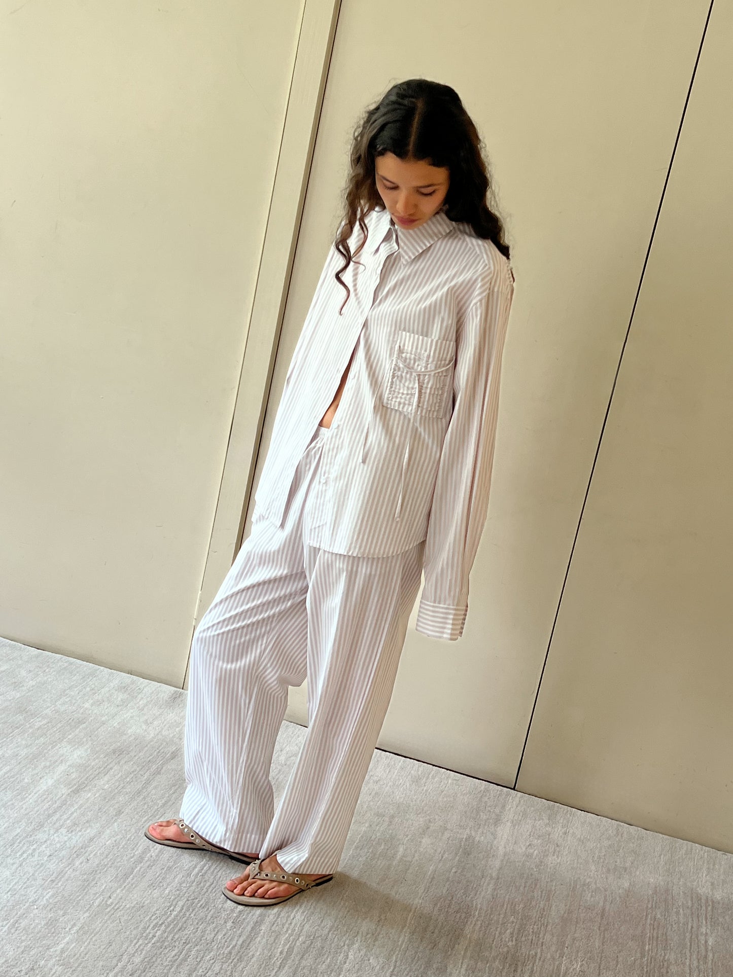 A woman with long, wavy hair stands indoors in the relaxed-fit Pocket Set - Lake Stripe by Deiji Studios and sandals, looking down with her hands by her sides against a neutral wall.