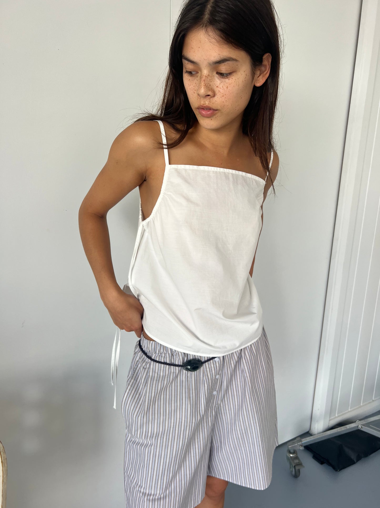 A young woman with long dark hair and freckles wears the Gathered Strap Top - White by Deiji Studios and loose, light blue striped shorts, standing indoors against a white wall and looking down to her right.
