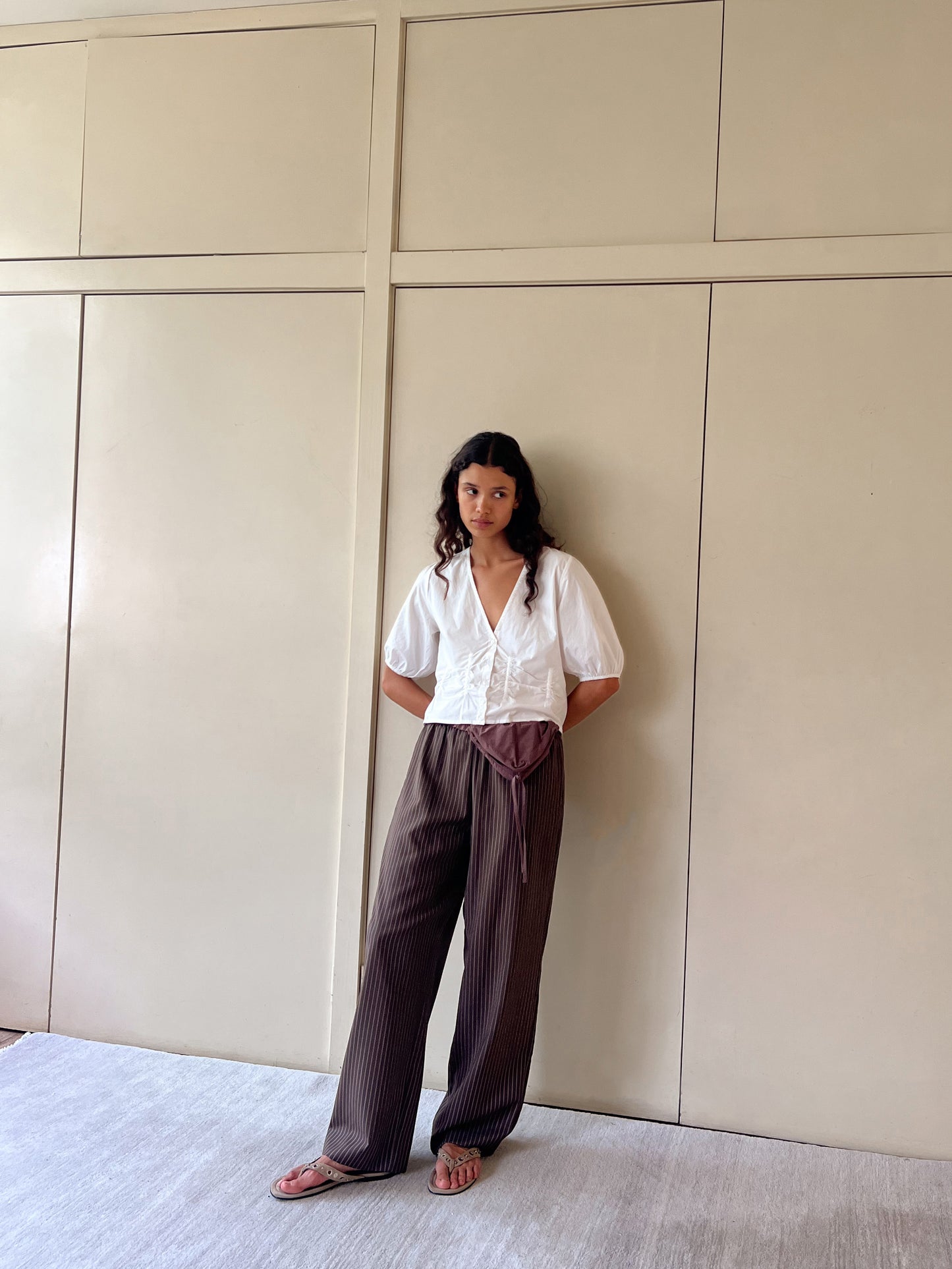 A woman with wavy hair and a relaxed posture stands against a beige paneled wall, wearing Deiji Studios Draw Top in white organic cotton poplin, brown striped wide-leg pants, a brown waist wrap accessory, and sandals.