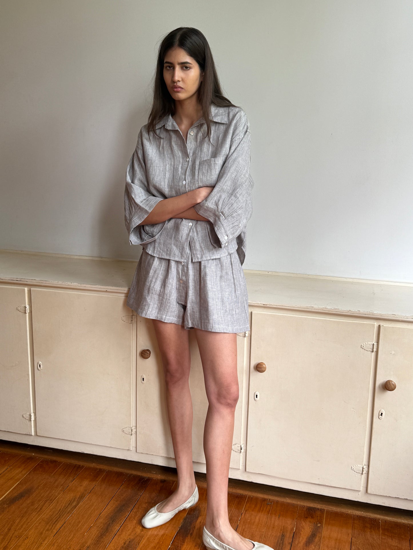 A woman with long dark hair stands indoors by a cream cabinet, wearing the 0 03 Set - Haze Blue made from GOTS certified organic, stone washed French linen. She pairs it with beige flats on a wooden floor in a minimal setting.