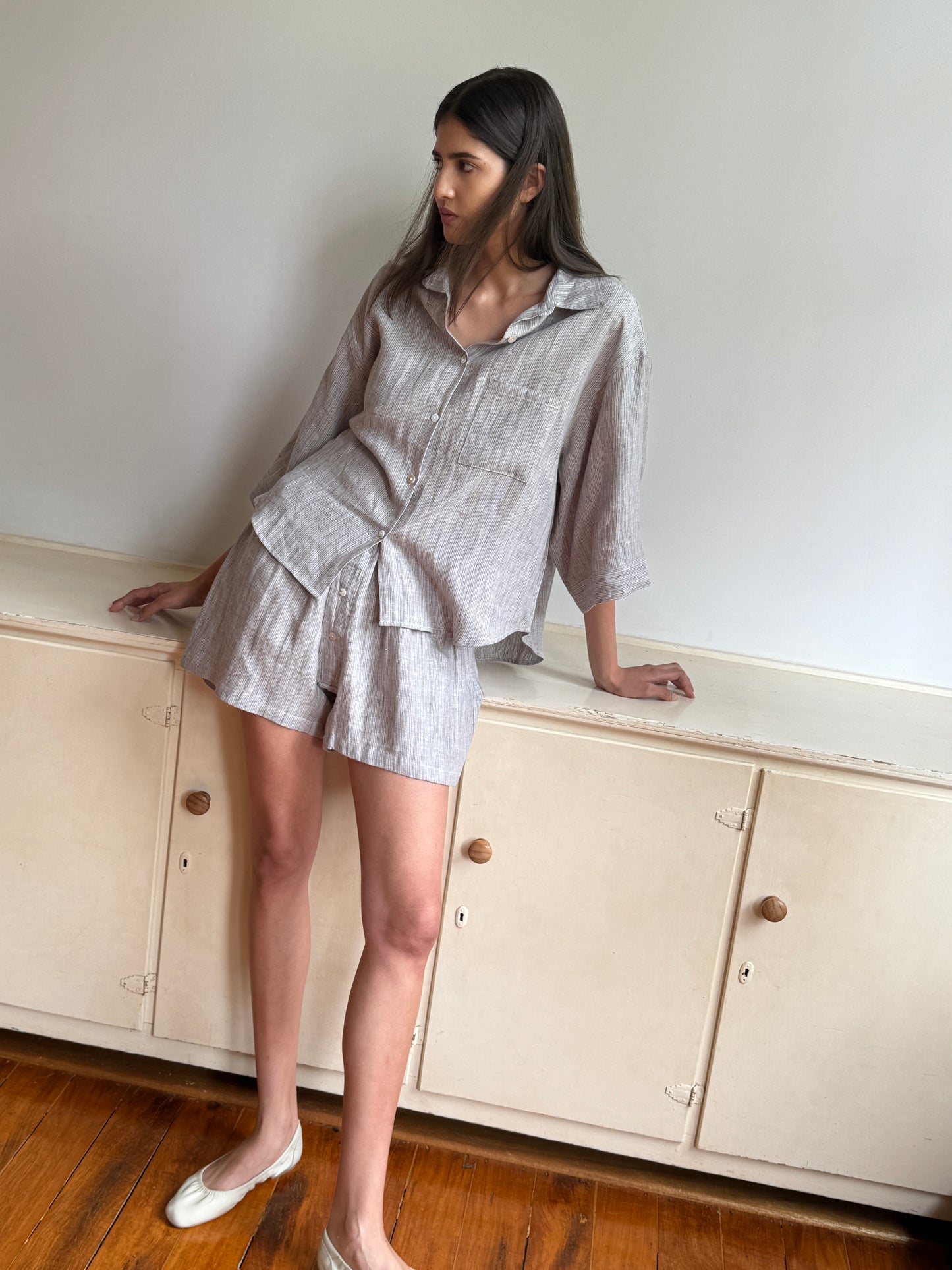 A woman with long dark hair wears the 03 Set - Haze Blue by 0, featuring a loose shirt and matching shorts. She leans against a cream cabinet on wooden flooring in a minimalist room with a white wall.