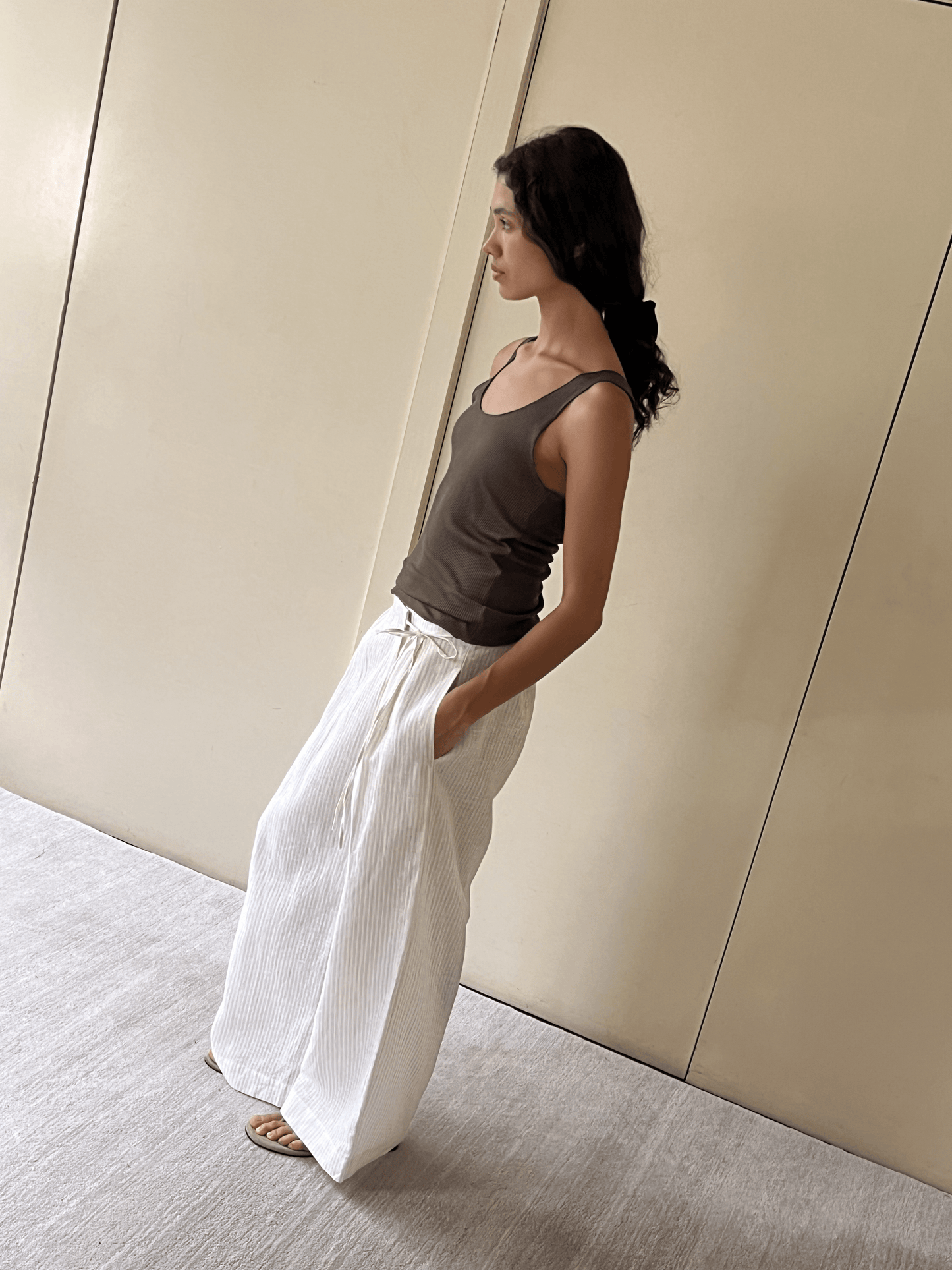 A woman stands indoors against beige panel walls, wearing the Deiji Studios Knit Tank in Stone with loose white pants and sandals. Her long dark hair is tied back with a ribbon, and she looks to the side with one hand in her pocket.