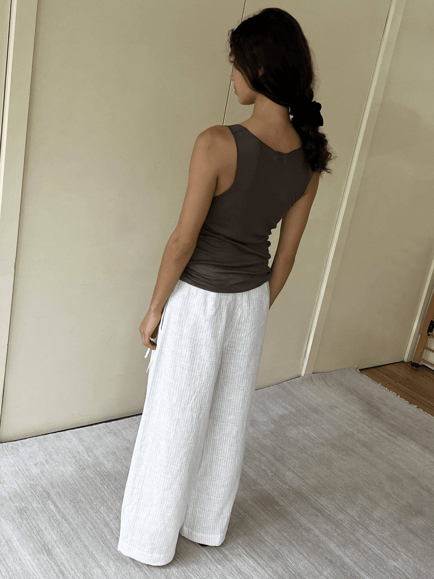 A person with long dark hair tied back wears the Knit Tank - Stone by Deiji Studios and loose white pants, standing indoors on a light carpet facing away from the camera.