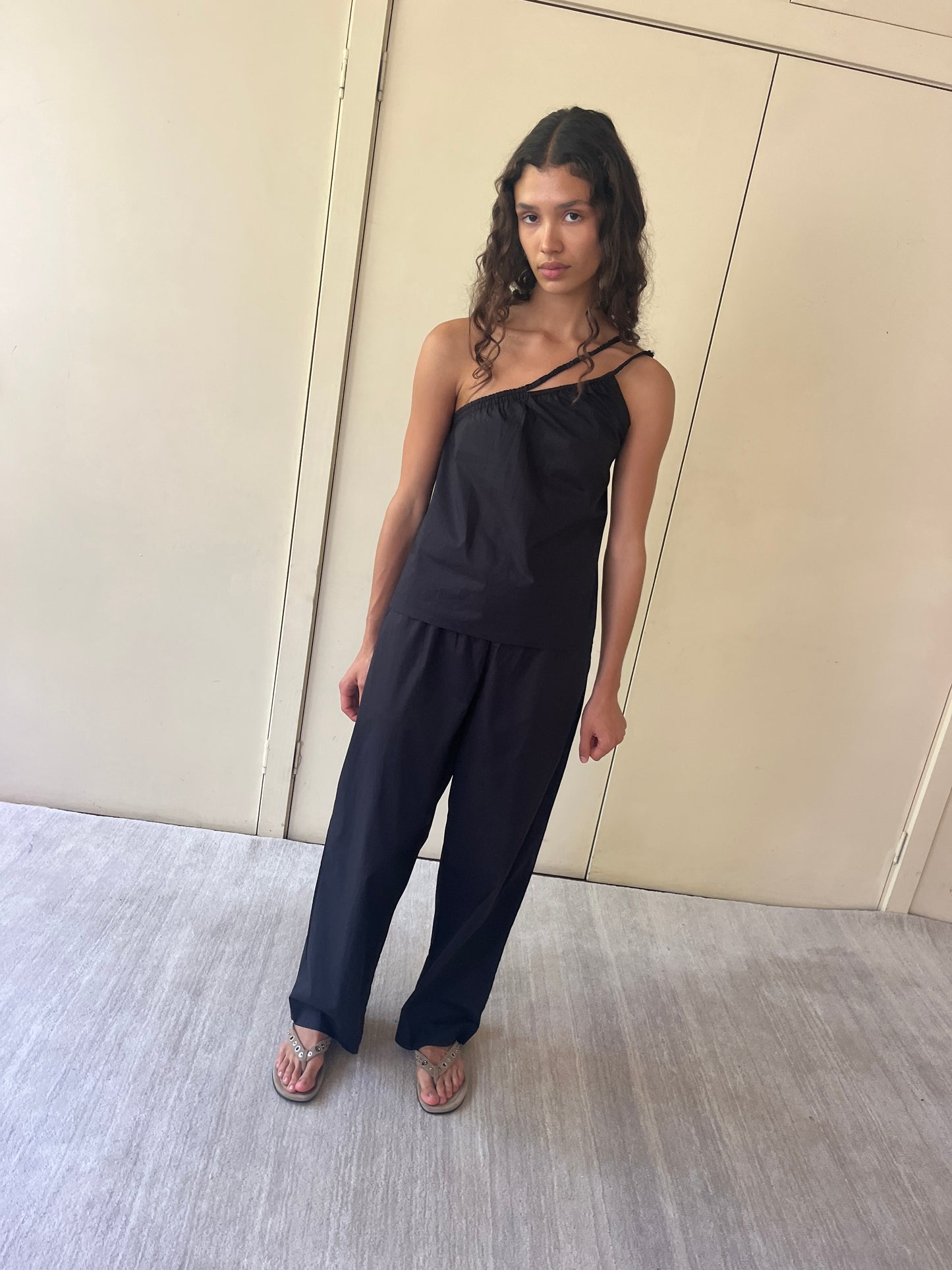 A woman with long, wavy hair stands indoors by a beige door, wearing the black Shoulder Top by Deiji Studios, loose organic cotton black pants, and sandals. She gazes at the camera with a neutral expression.