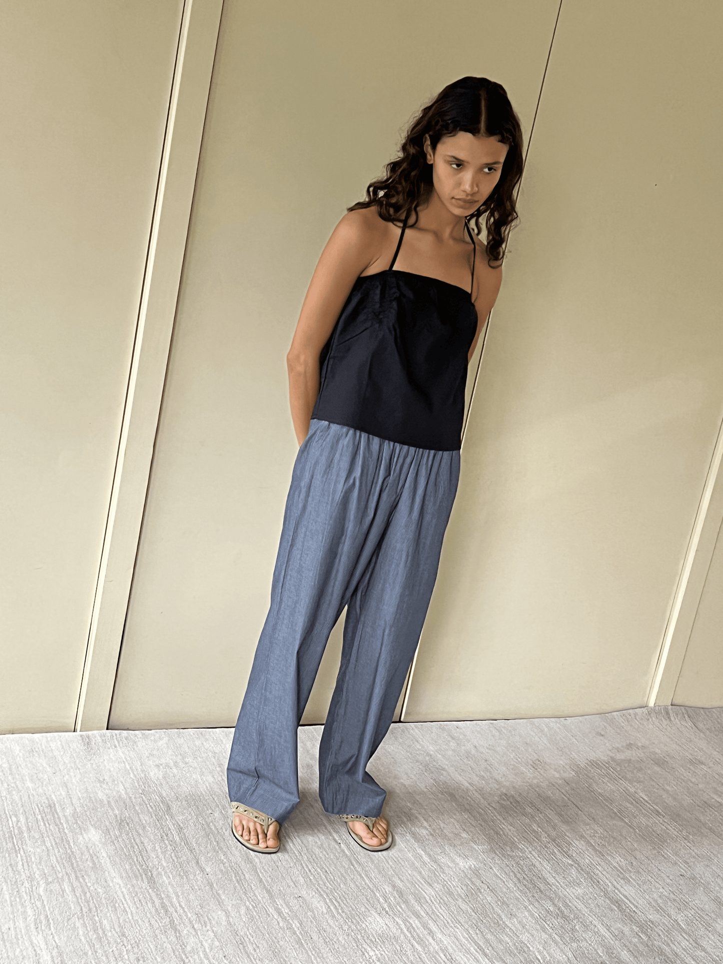 A woman with wavy brown hair stands indoors against a beige wall, wearing the Ease Trouser in Blue Pinstripe—relaxed organic cotton pants with an elastic waist—paired with a black halter top and sandals, looking slightly down.