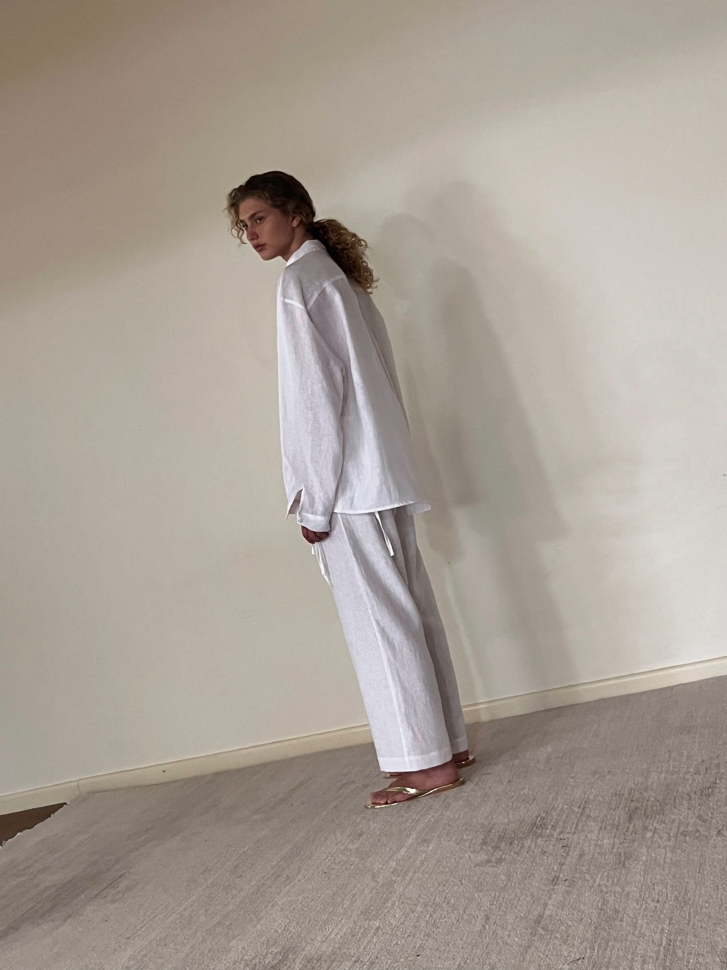 A person with curly hair stands sideways in a minimalist room, barefoot on a light carpet, wearing the String Linen Set - White against a plain wall.