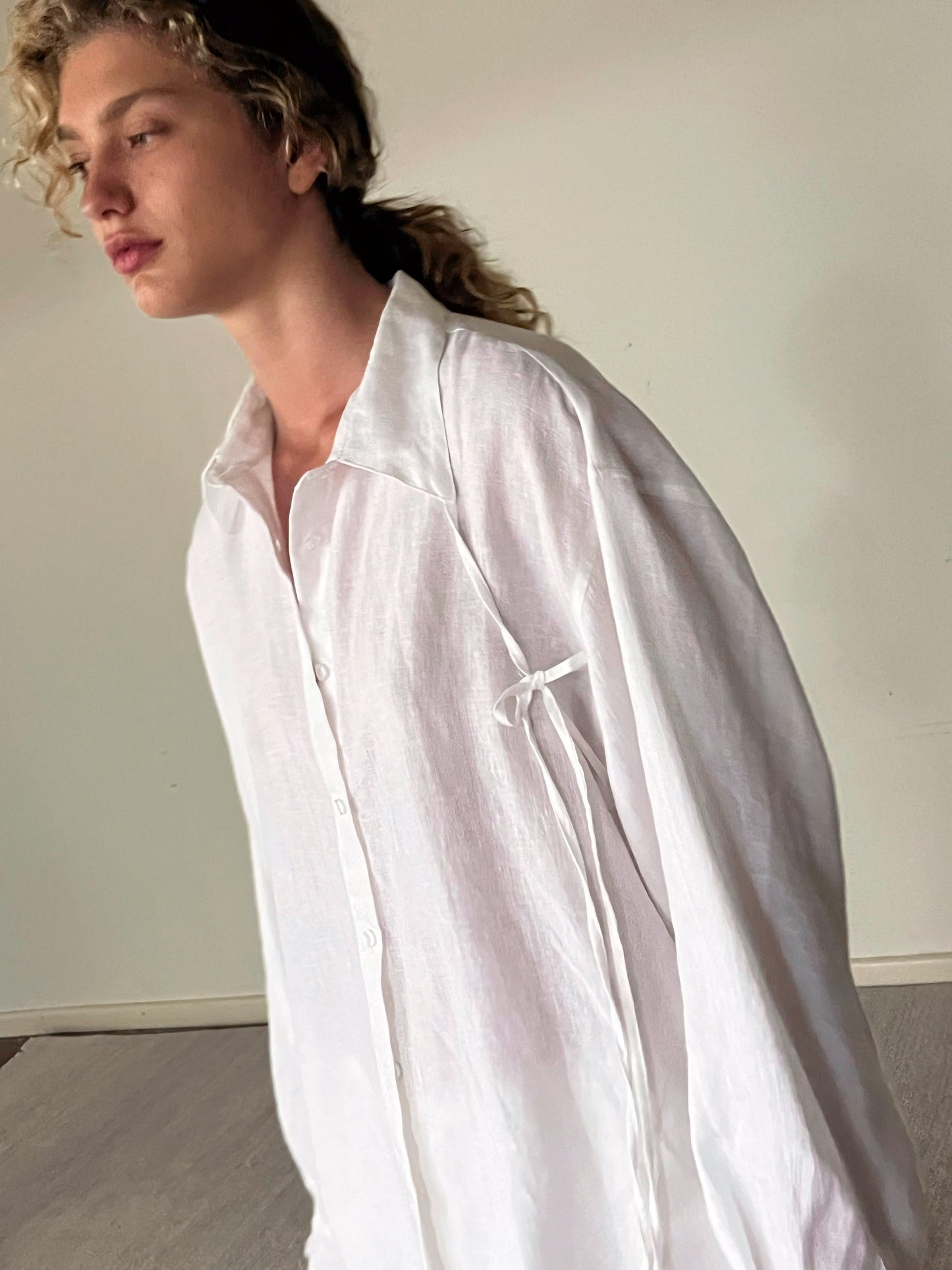 A person with curly blonde hair wears the String Linen Set - White, standing indoors against a plain wall and looking to the left with a calm expression.