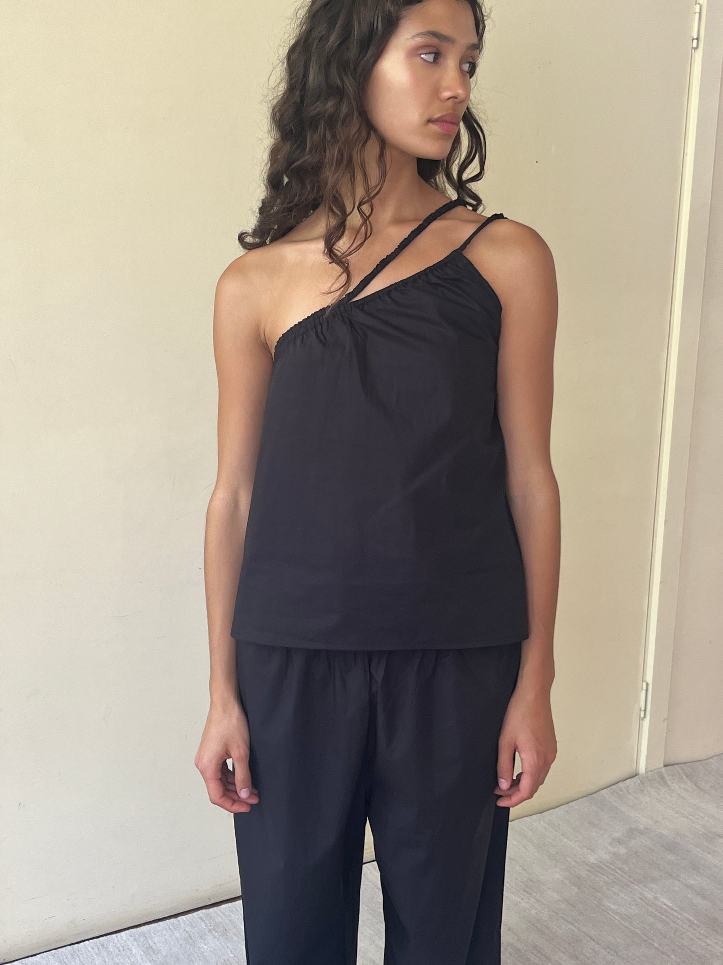 A woman with wavy brown hair stands indoors against a beige wall, looking left, wearing a sleeveless, one-shoulder black Shoulder Top - Black and matching loose organic cotton pants.