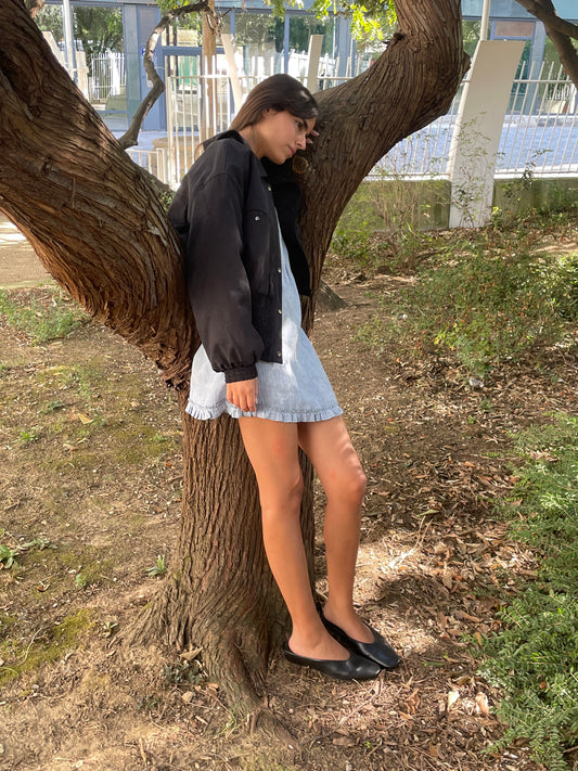 A young woman in a Deiji Studios Shirring Jacket - Slate, light denim dress, and black shoes leans against a tree outdoors, one leg bent and eyes closed amid grass and foliage.