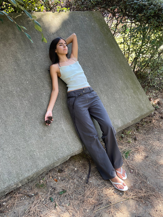 A woman with long dark hair, dressed in the Fine Knot Tank - Ice Blue, gray tailored fit pants, and white sandals, lounges on a slanted concrete surface outdoors among greenery. She holds pine cones in one hand and looks relaxed.