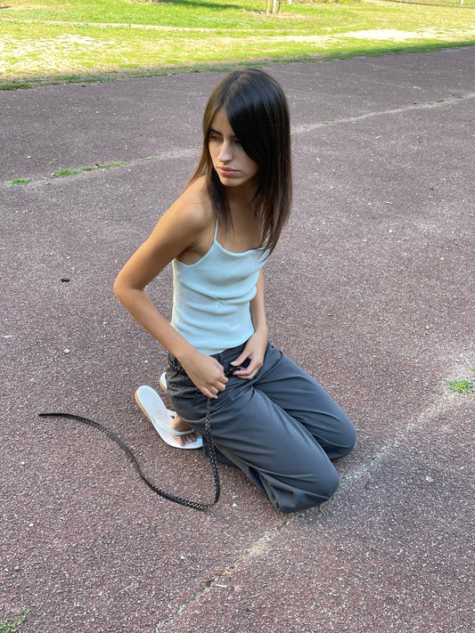 A young person with long dark hair kneels on an outdoor track, holding a black rope. They wear a light blue tank top, Zip Pant - Charcoal, and white sandals. Grass and trees are visible in the background.