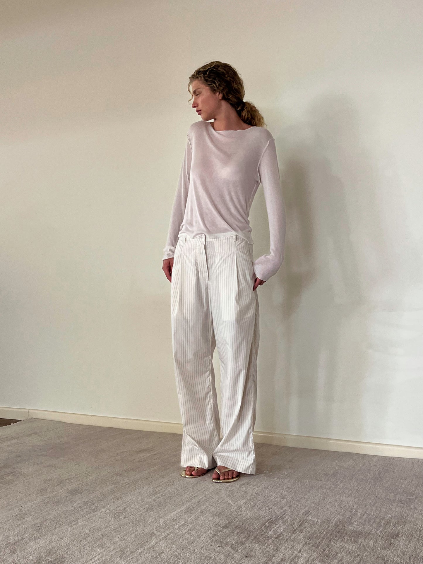 A person with curly hair stands indoors, wearing the Knit Long Sleeve - Off White top and loose, high-waisted white pinstripe pants with sandals. The outfit features a responsible blend for OEKO-TEX 100 certification.