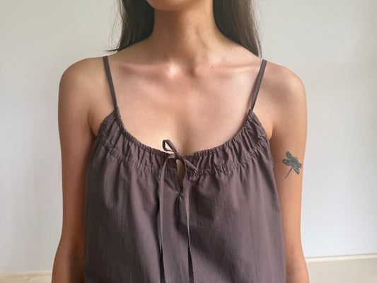 A woman in the Light Dress, a sleeveless brown piece with thin straps, stands against a plain background. A dragonfly tattoo decorates her upper left arm while her face is partially out of frame.