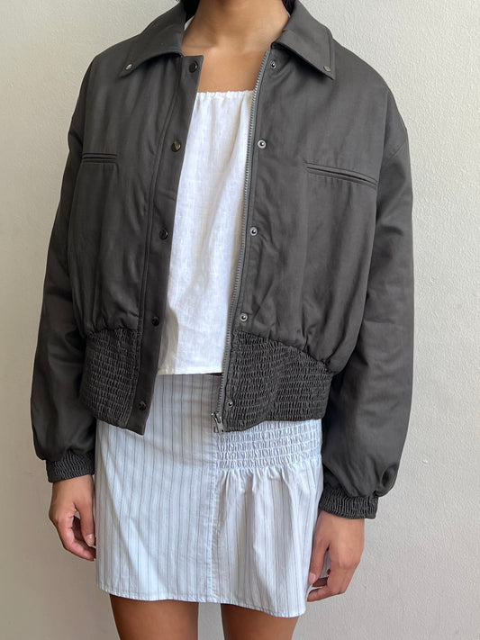 A person wears the Shirring Jacket - Slate by Deiji Studios, featuring ruched cuffs and hem, over a white sleeveless square-neck top and a white skirt with light blue vertical stripes, standing against a plain light-colored wall.