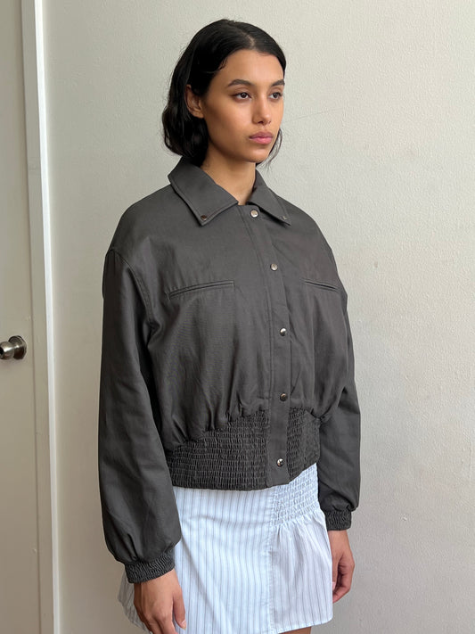 A person with medium-length dark hair wears the Shirring Jacket - Slate by Deiji Studios, featuring front pockets, paired with a light blue and white striped skirt, standing against a plain light-colored wall.