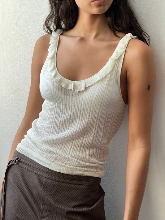 A woman wearing the Deiji Studios Neck Ruffle Knit - Off White and brown pants stands against a light-colored wall, her face partially out of frame.