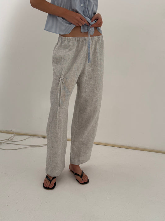 A person wears the Eyelet Pant - Fine Stripe, ankle length and relaxed, with a light blue shirt tied at the waist and black sandals, standing on a white floor against a plain wall—Deiji Studios-inspired style.