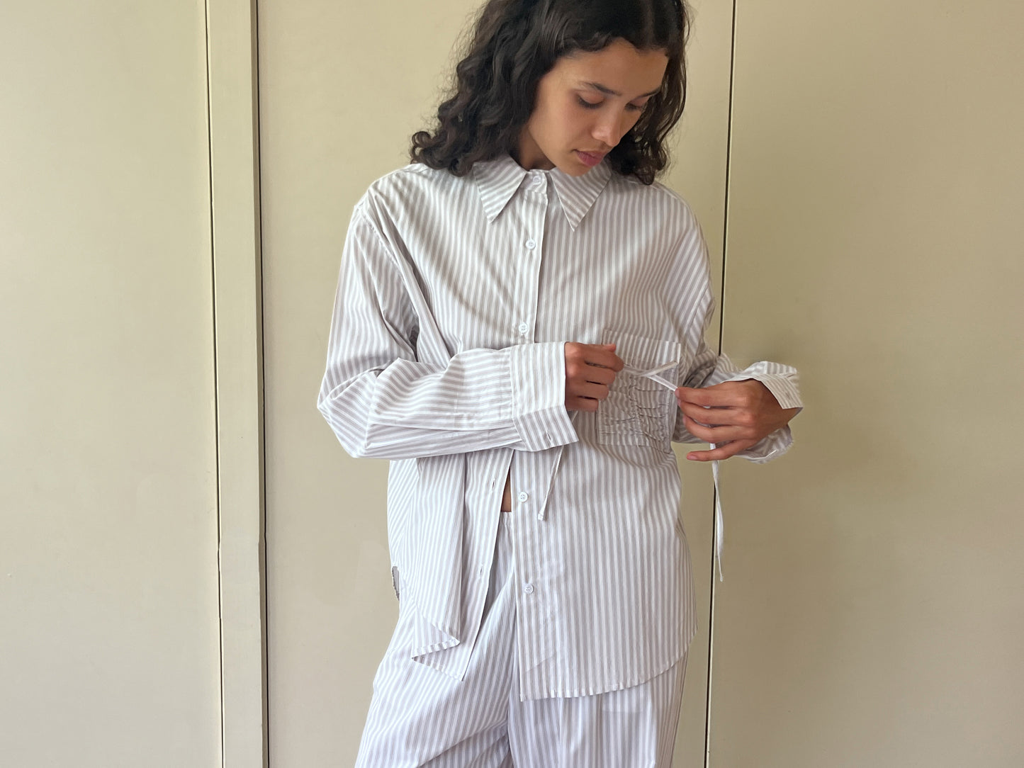 A woman with long curly hair wears the Pocket Set - Lake Stripe by Deiji Studios, a relaxed, light-striped button-up shirt and matching pants, indoors while looking down and adjusting or examining the shirt’s pocket or cuff.