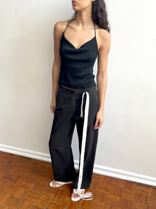 A woman stands indoors against a white wall wearing the Fine Knot Tank - Black from Deiji Studios, loose black pants with a long white rope belt, and white sandals. She has long wavy hair and looks slightly to the side.