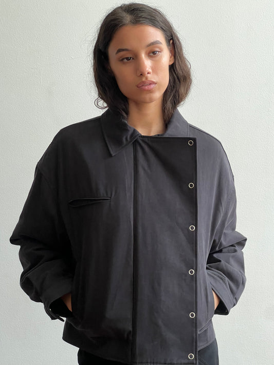 A person with shoulder-length dark hair stands against a plain light wall, wearing the Lapel Jacket - Black from Deiji Studios featuring a high collar and snap buttons, hands in pockets and a neutral expression.