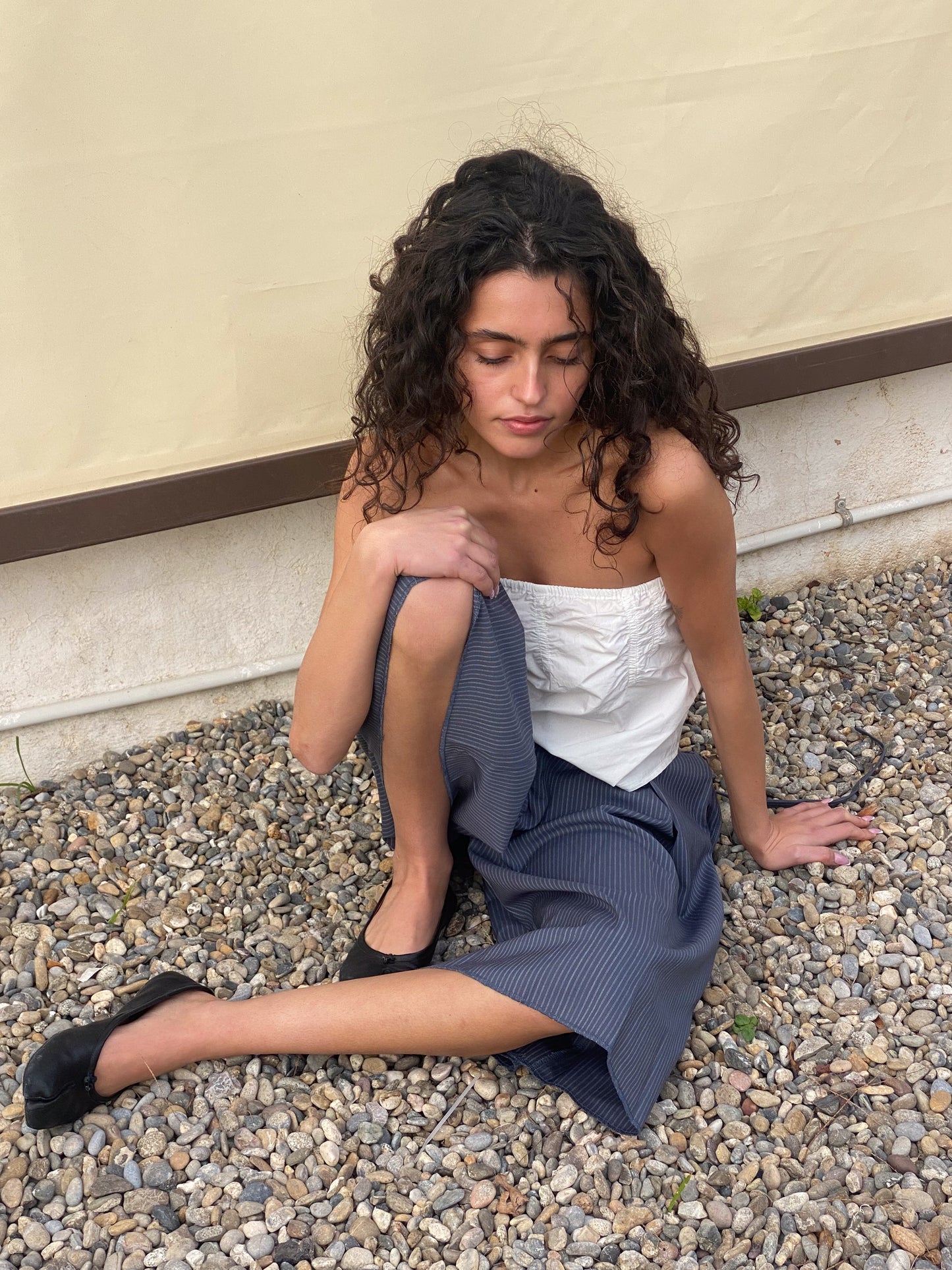 A woman with curly hair wears the Tube Top - White by Deiji Studios and a blue skirt, sitting on gravel with one knee up and an arm resting on it beside a beige wall—a serene, thoughtful scene.