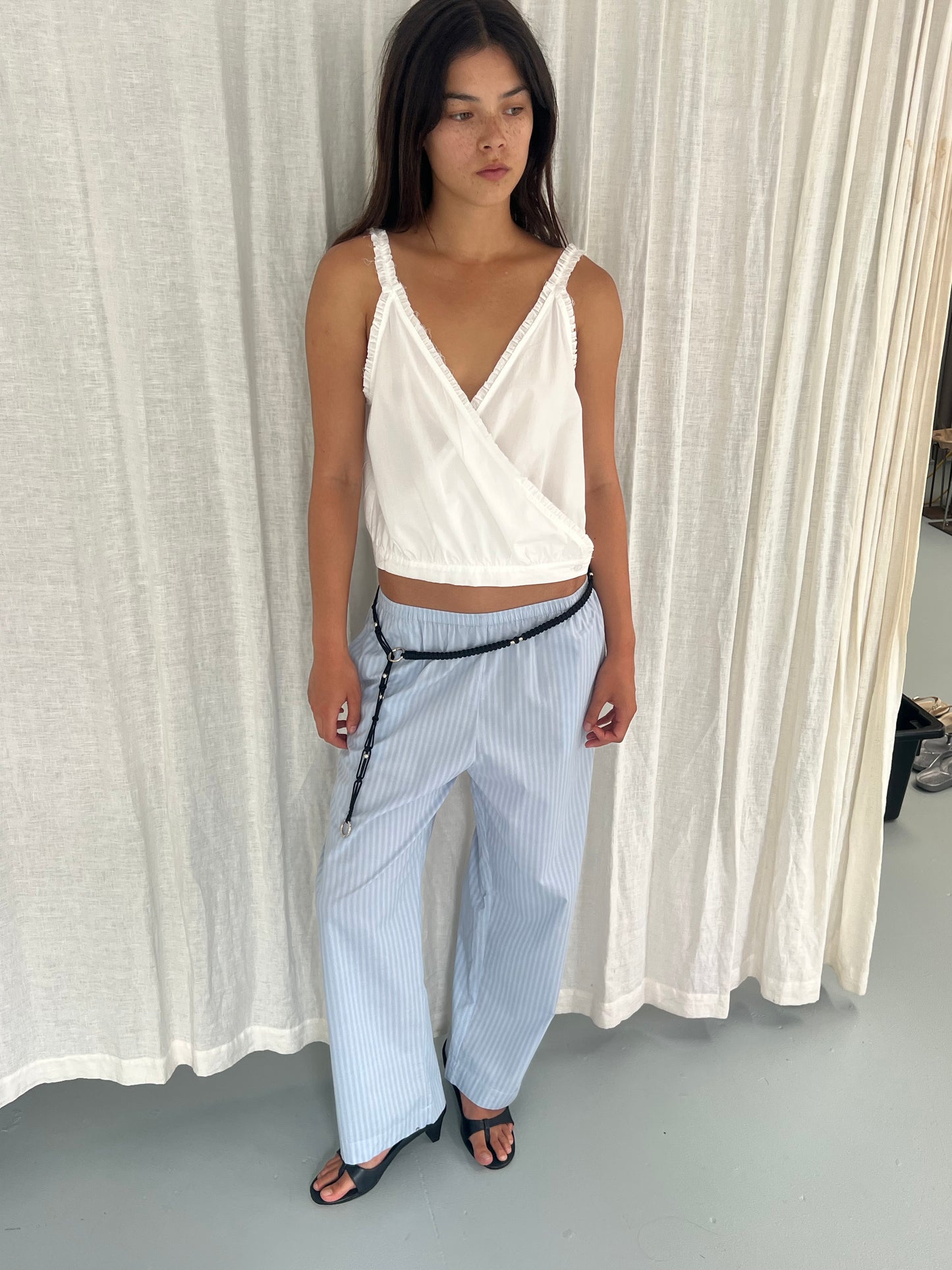A woman stands indoors in a sleeveless white wrap top, paired with the Ease Trouser - Lake Blue Stripe by Deiji Studios, black sandals, and a thin black belt. White curtains are visible in the background.